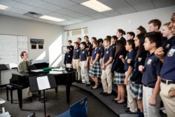 A group of students in music class listen to the piano